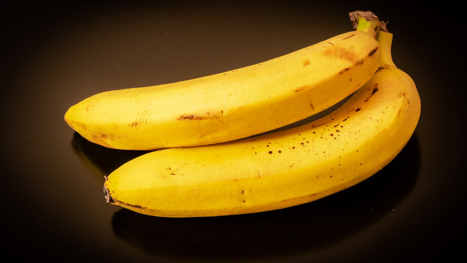 Două banane galbene cu pete brune, pe fond negru, fotografiate de Krzysztof Golik