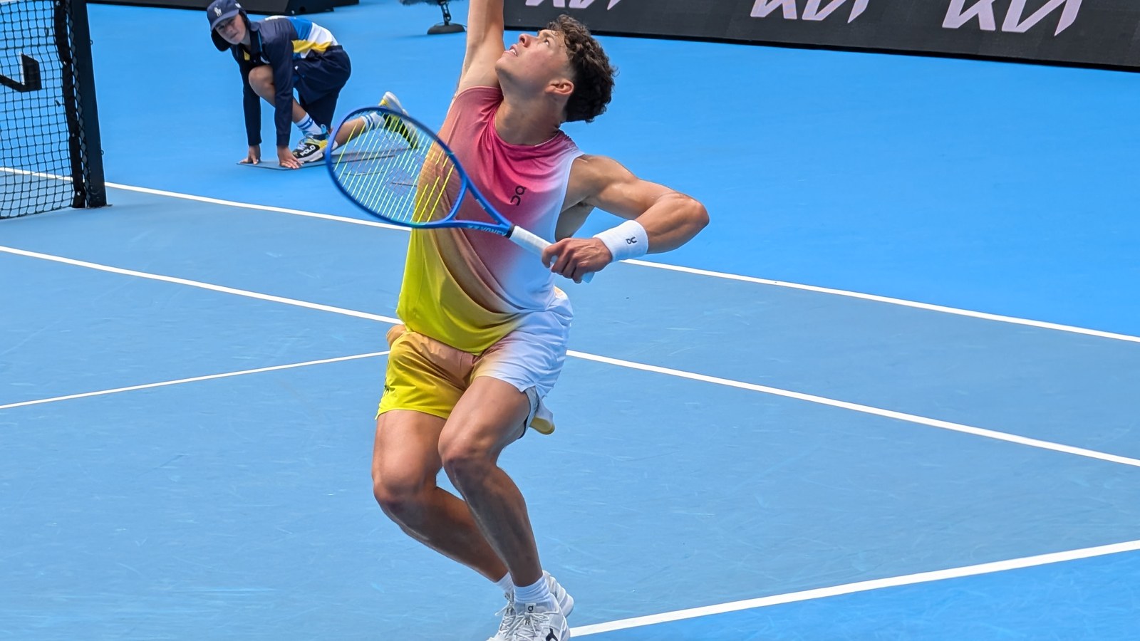 Ben Shelton la serviciu pe un teren albastru de hard, cu rachetă albastră și echipament roz-galben, la Australian Open 2…