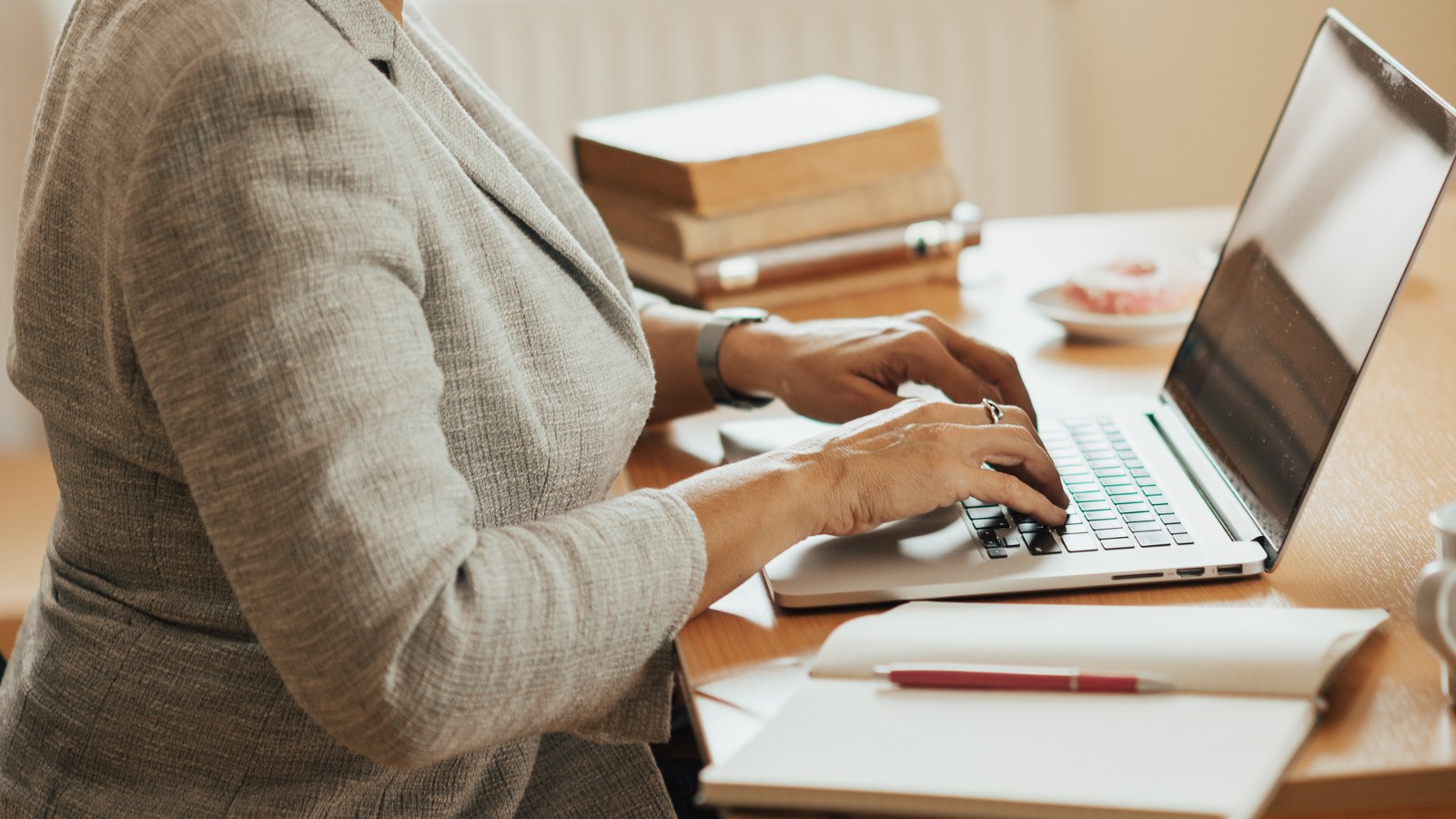 Persoană care lucrează la un laptop, cu un caiet deschis pe birou